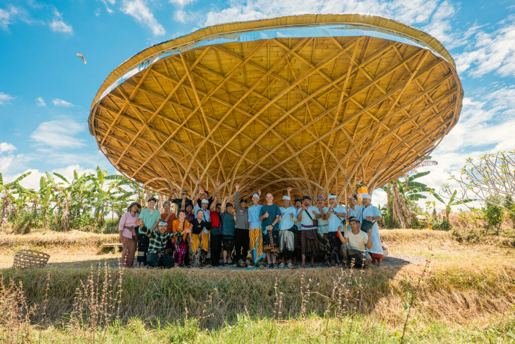 Group of people posing under a large woven bamboo canopy outdoors on a sunny day, with blue sky above.